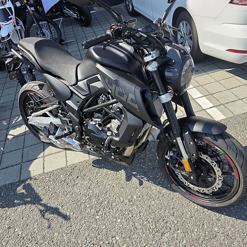 Ein schwarzer Motorrad mit dem Wort 'HCL' auf der Seite ist auf einem gepflasterten Bereich neben einem weißen Auto geparkt.