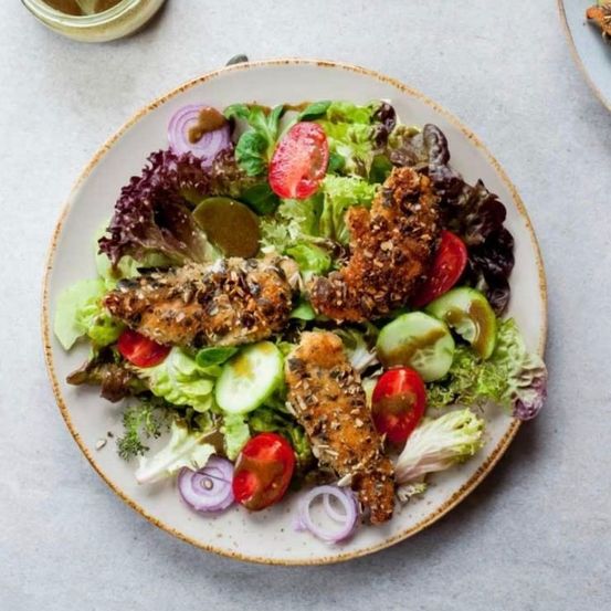 Ein Teller mit Salat, Tomaten, Gurken und paniertem Hähnchen. Der Salat besteht aus Salat, roter Zwiebel und Dressing.