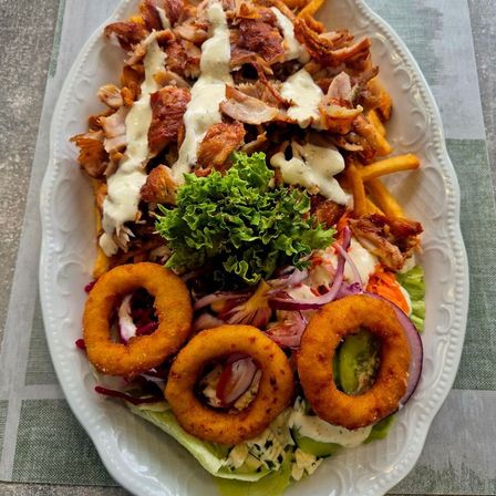 Ein weißer Teller mit einem Fleischgericht mit geshreddetetem Huhn, Pommes und Zwiebelringen. Salat, rote Zwiebeln und andere Gemüsesorten sind auf dem Teller.