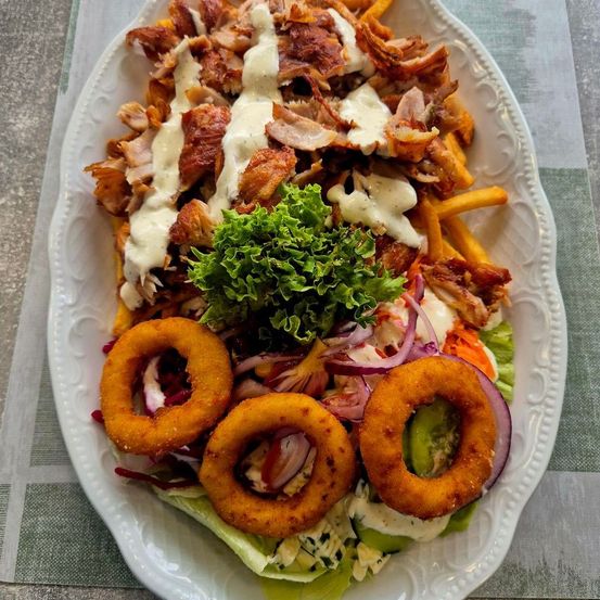 Ein weißer Teller mit einem Fleischgericht mit geshreddetetem Huhn, Pommes und Zwiebelringen. Salat, rote Zwiebeln und andere Gemüsesorten sind auf dem Teller.