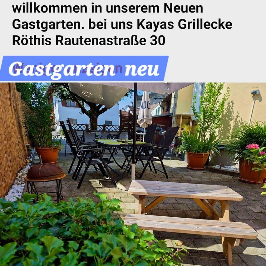 A garden area with a wooden table and chairs, a fire pit, and plants. It has a brick floor and is shaded by an umbrella.