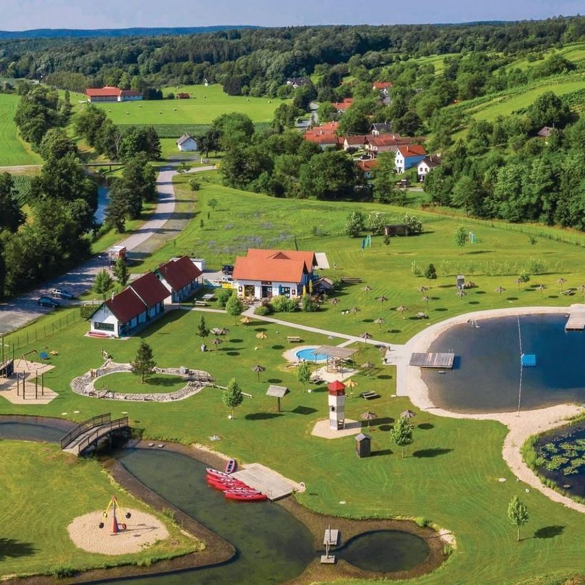 Ein Luftbild einer ländlichen Gegend mit mehreren Häusern, einem See und einem Spielplatz. Bäume und grüne Felder umgeben das Gebiet, und es gibt eine Brücke und einen kleinen Steg.