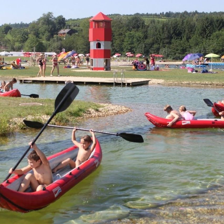 Zwei Kinder paddeln ein rotes aufblasbares Boot. In der Nähe schwimmen und entspannen sich Menschen am Ufer mit Schirmen und einem Rettungsturm.