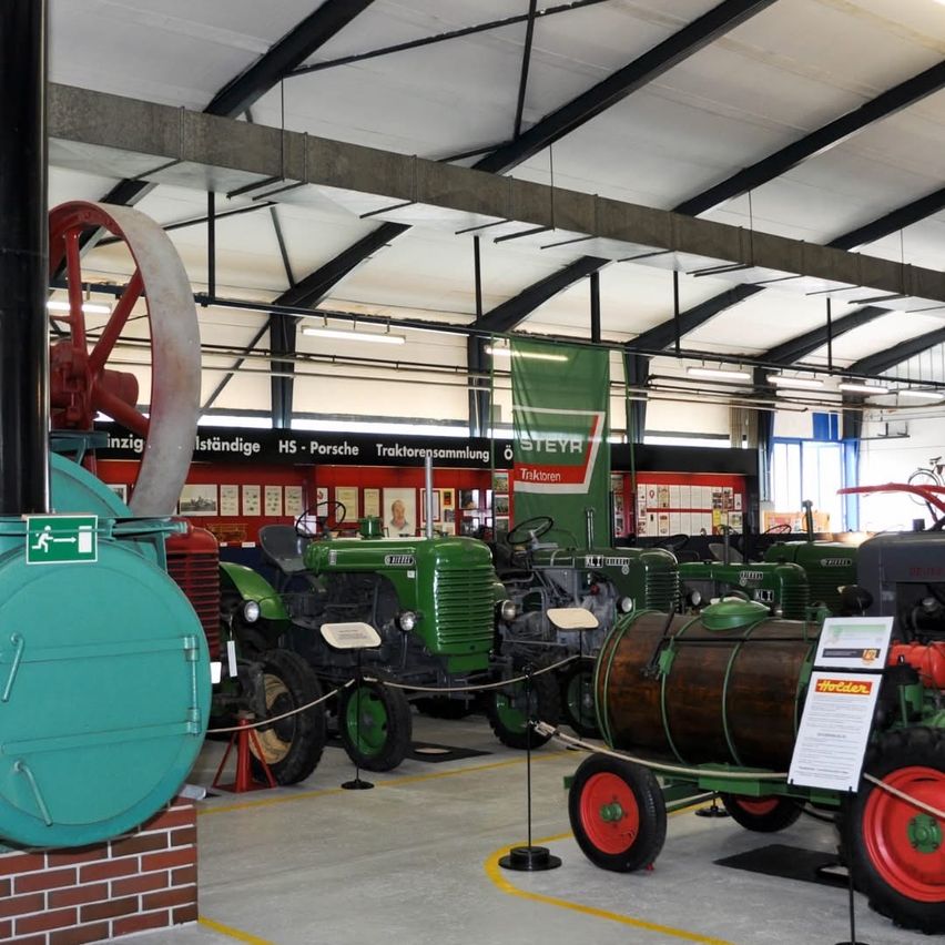 Ein Lagerhaus zeigt mehrere alte grüne Traktoren. Ein rotes Rad ist prominent auf der linken Seite ausgestellt. Die Decke hat Stahlträger und Leuchten.