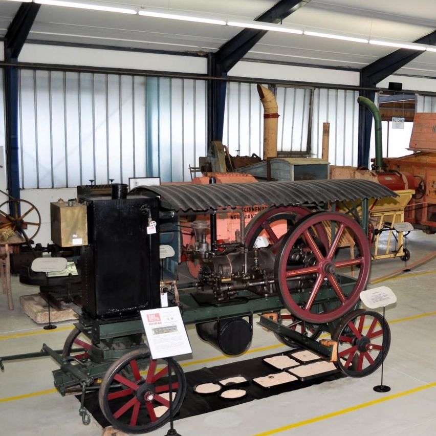 Ein altes, schwarzes Dampffahrzeug mit großen roten Rädern ist in einem großen Innenraum ausgestellt, umgeben von anderen alten Maschinen.