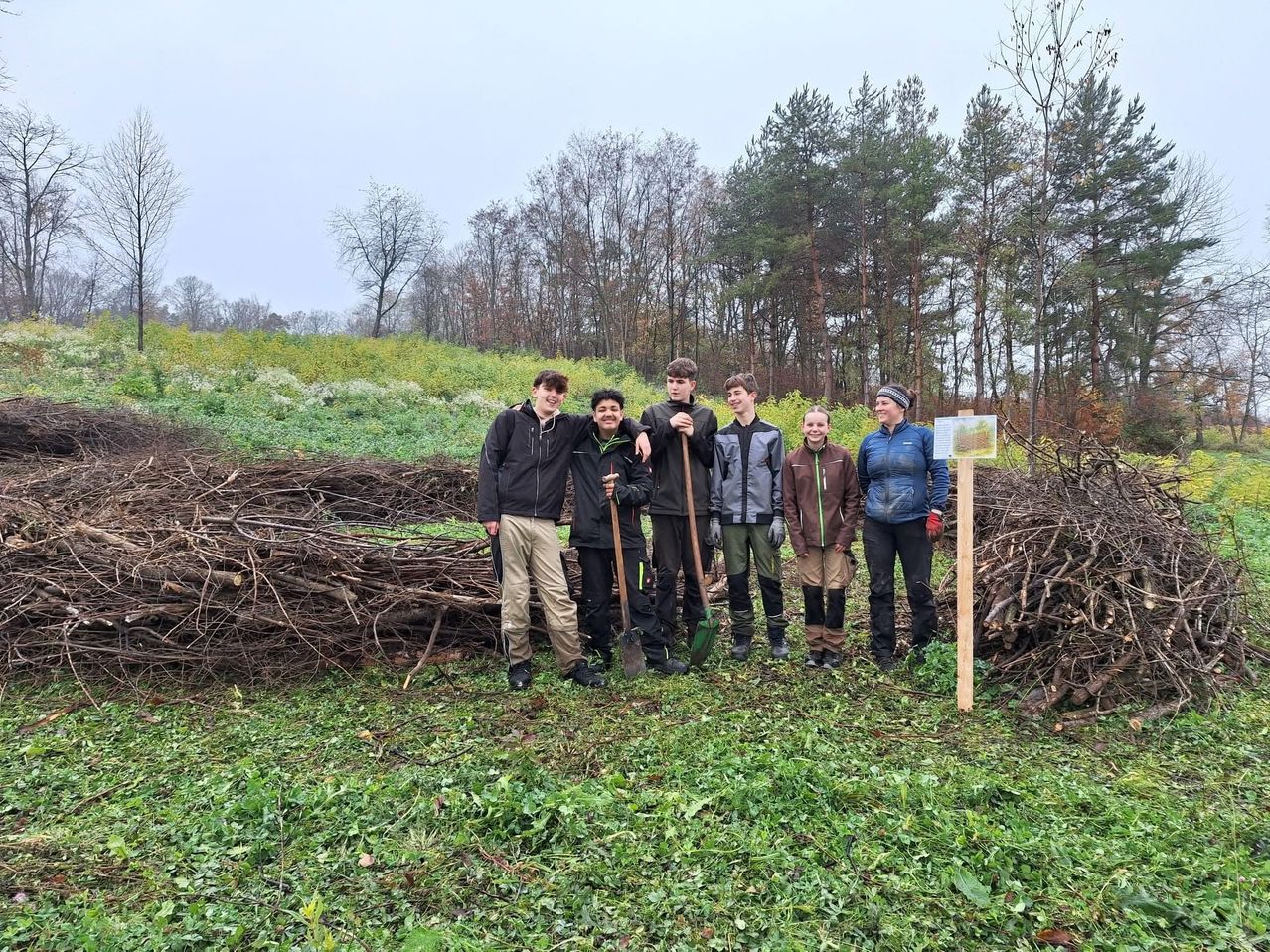Five individuals, including two women and three men, stand in a grassy field, holding shovels and a rake. Behind them, a pile of sticks is present. In the background, a dense forest is visible.