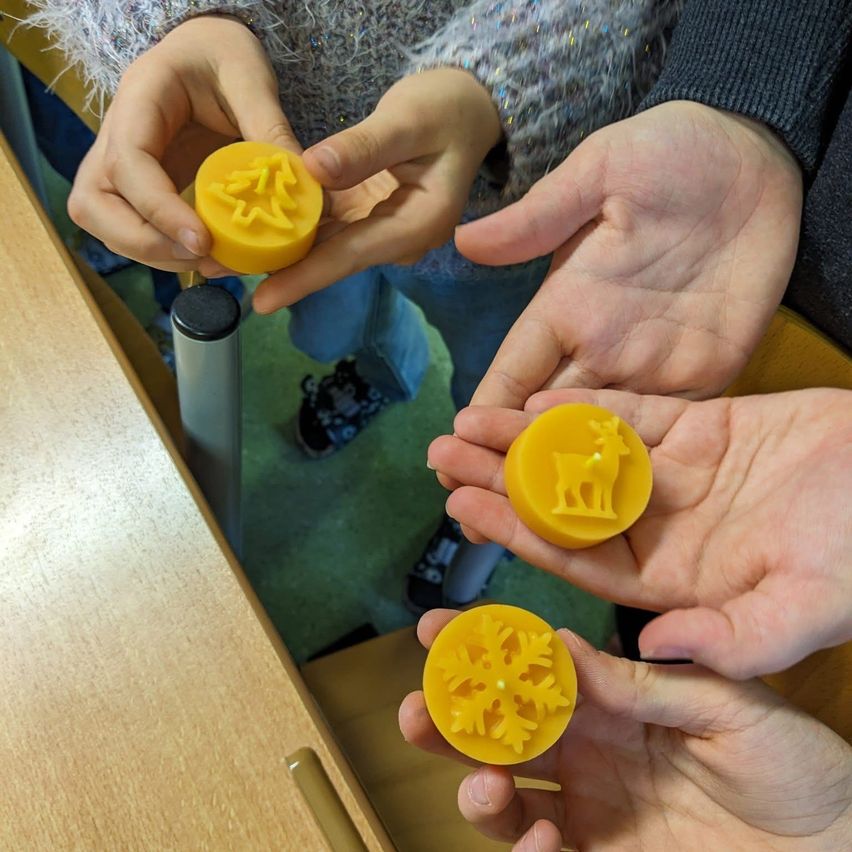 Three hands hold yellow candles with winter designs, likely during a holiday craft session. The table is wooden, and the floor is carpeted.