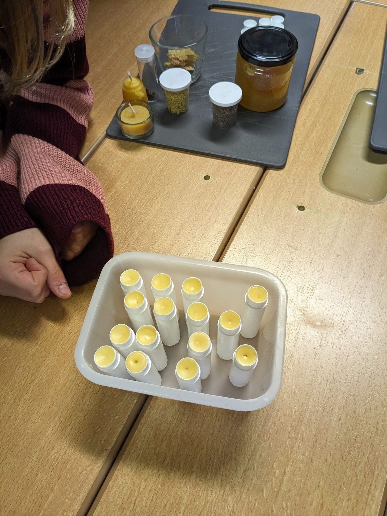 A person sits at a wooden table, with a tray of small containers and a tray of lip balms in front of them. The lip balms are in a white plastic container, some are open.