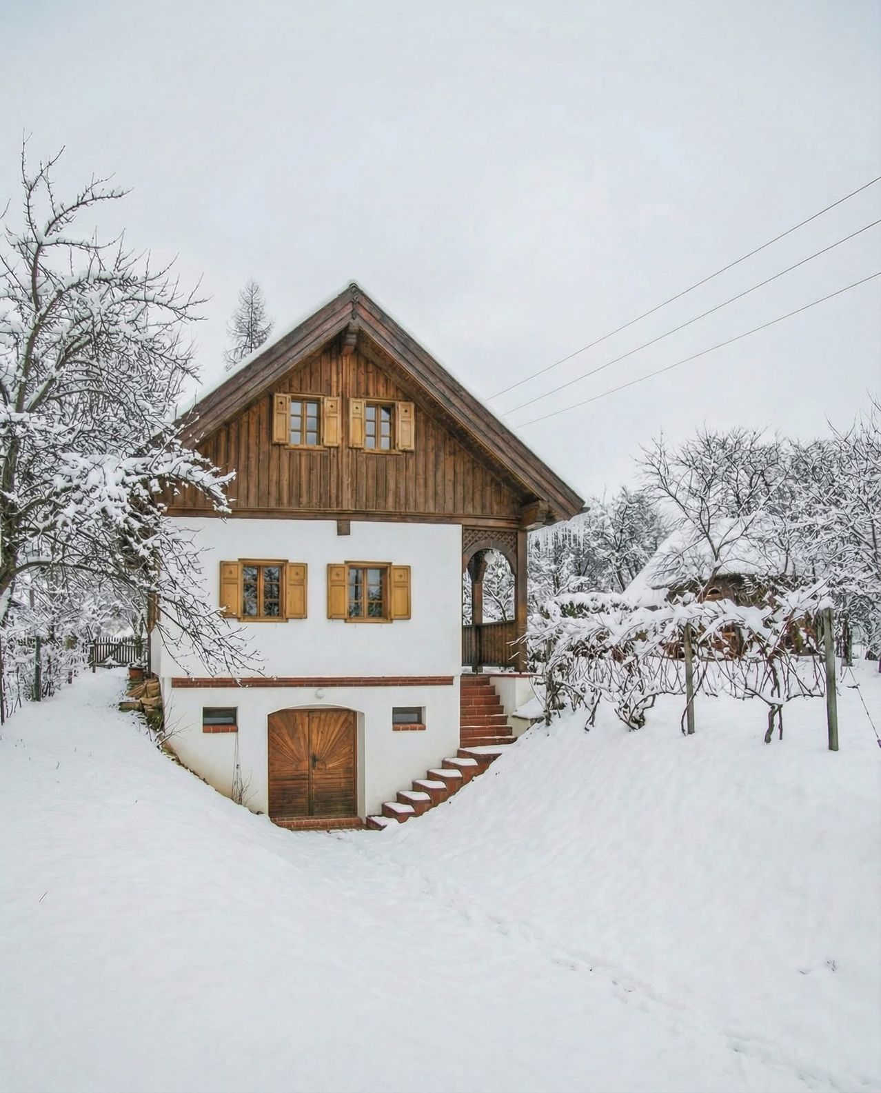 Ein kleines, weißes Haus mit braunen Holzakzenten, bedeckt mit Schnee, mit einer Holztür, Stufen und einer Veranda. Es ist von schneebedeckten Bäumen und Büschen umgeben.