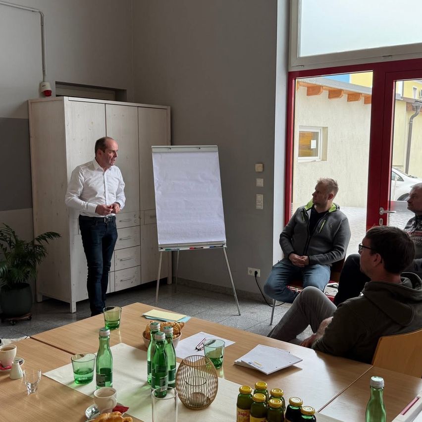 Ein Mann steht an einer Tafel und spricht zu drei sitzenden Männern. Der Raum hat einen Tisch mit Gläsern und Essen, einen Topf und eine Glastür.