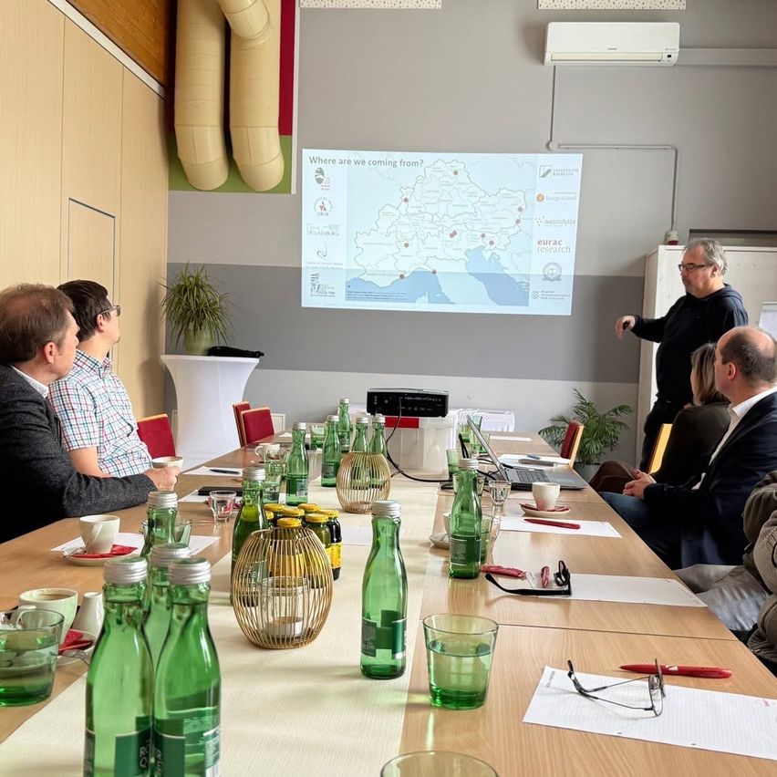 Eine Gruppe von Menschen sitzt in einem Besprechungsraum an einem Tisch. Ein Mann steht vor einer Projektionsleinwand mit einer Karte. Flaschen und Papiere liegen auf dem Tisch.