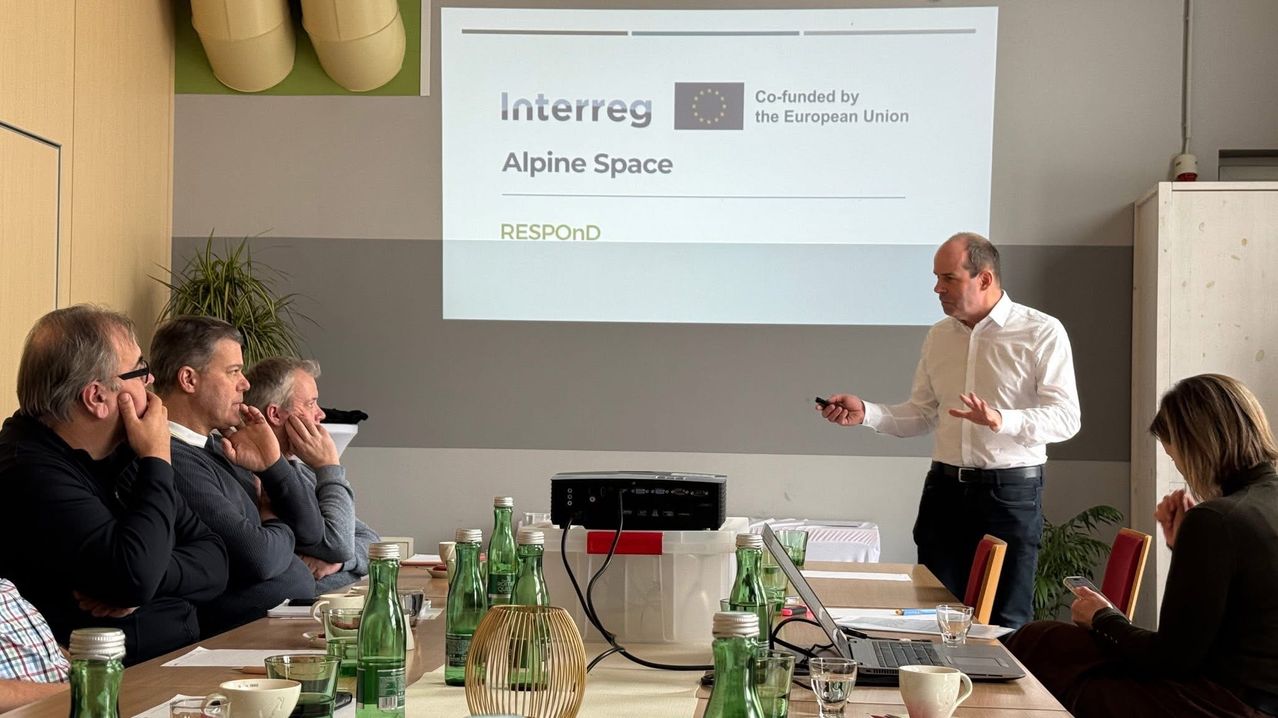 Eine Präsentation wird in einem Konferenzraum gehalten. Ein Mann steht vor einer Projektionsleinwand. Ein Tisch mit Flaschen und einem Laptop steht im Vordergrund.