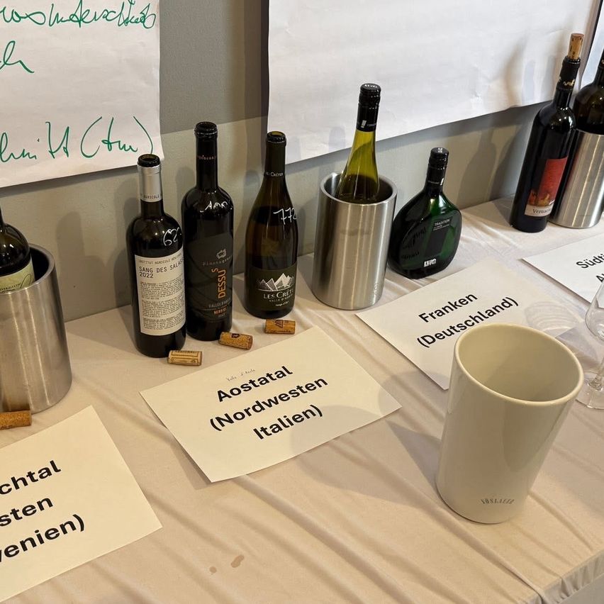 Ein Tisch mit mehreren Weinflaschen, die mit Aostatal, Nordwest-Italien und Fränkische Region beschriftet sind. Die Flaschen sind in silbernen Eimern aufbewahrt und werden von Weingläsern und Schildern begleitet.