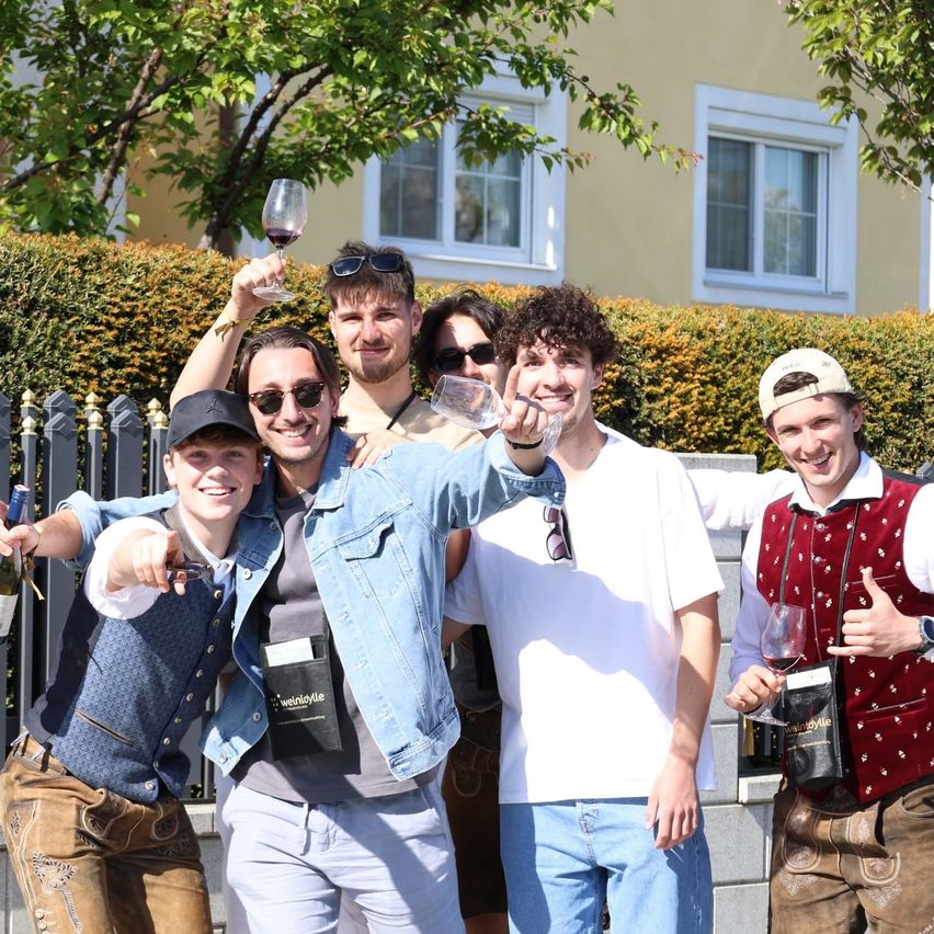 Sechs Männer stehen im Freien, lächeln und posieren für ein Foto. Sie halten Weingläser und eine Flasche. Dahinter steht ein Busch und ein Haus mit weißen Fenstern.