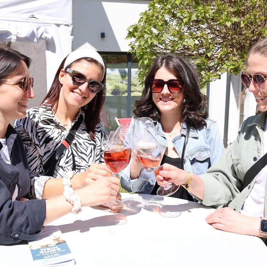 Vier Frauen lächeln und heben Weinglaser, möglicherweise bei einer Outdoor-Veranstaltung. Sie tragen Sonnenbrillen und Jacken. Ein Buch mit dem Titel 'Wein Guide' liegt auf dem Tisch.