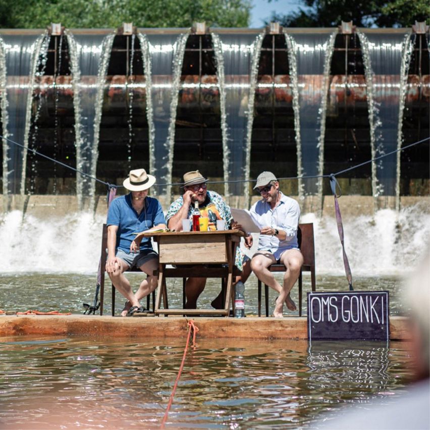 Drei Männer sitzen auf einer Holzplattform in einem Fluss, mit einem Damm und fließendem Wasser hinter ihnen. Sie tragen Hüte und scheinen in ein Gespräch vertieft zu sein.