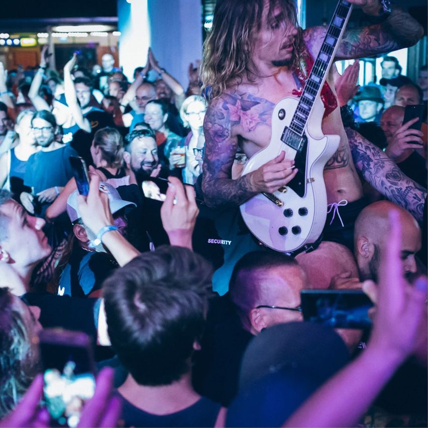 Eine Rockband tritt bei einem Konzert auf der Bühne auf, der Leadsänger ist oberkörperfrei und hält eine Gitarre, während das Publikum jubelt und Fotos macht.