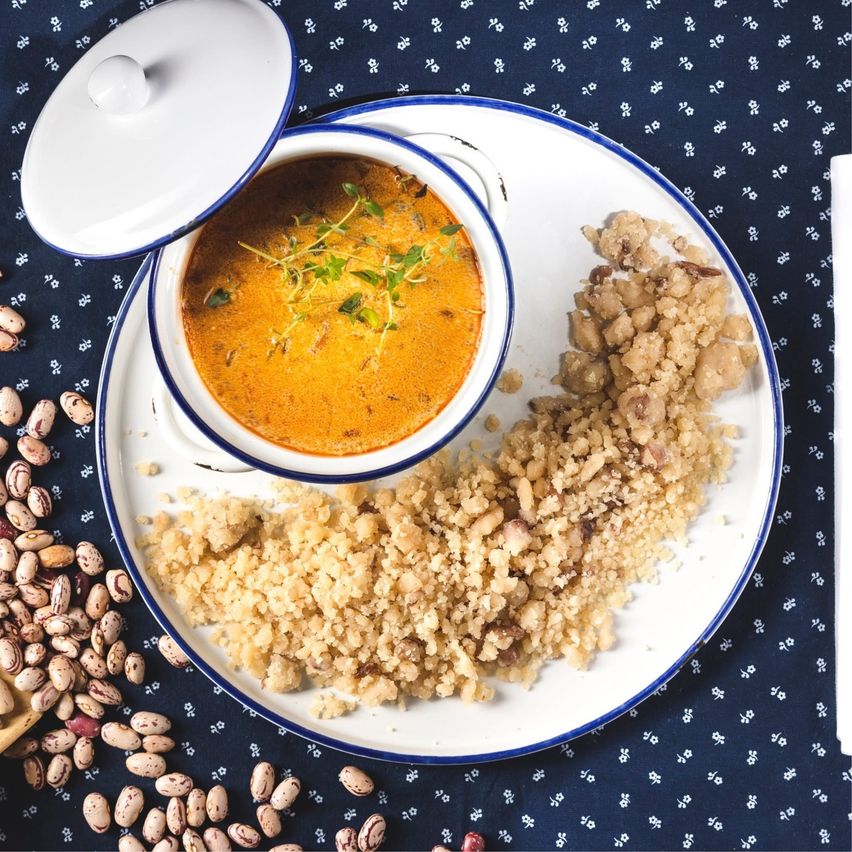Ein weißer Teller mit blauem Rand hält eine Schüssel mit orangefarbener Suppe mit Kräutern, einer krümelige Beilage und Bohnen auf der Seite.
