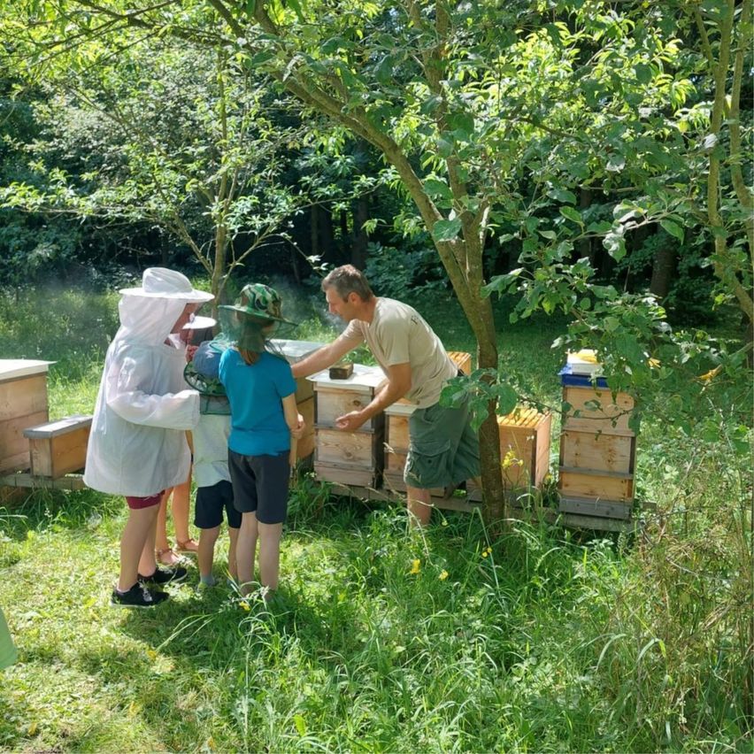 A beekeeper wearing a white suit is standing next to a man in a forest, with a group of children observing.