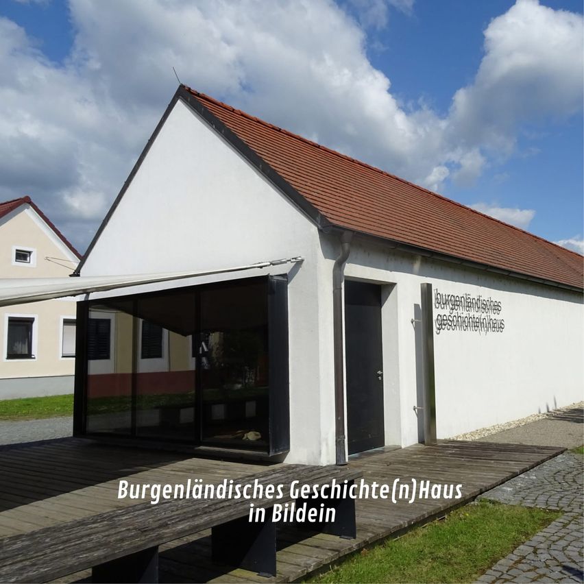 Ein weißes Haus mit braunem Dach, beschriftet mit Burgenland Geschichte Haus in Bildein, steht auf einem Kopfsteinpflasterweg. Es hat Glasfenster und eine schwarze Tür. Vorne steht eine Bank.