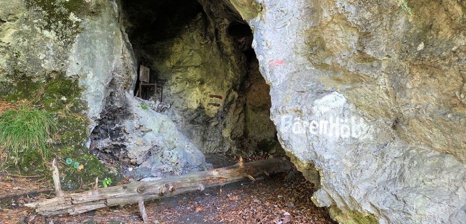 Ein Höhleneingang mit einem Holzstamm, Moos und einem Stuhl im Inneren. Die Felswand hat weiße Farbe mit Text.