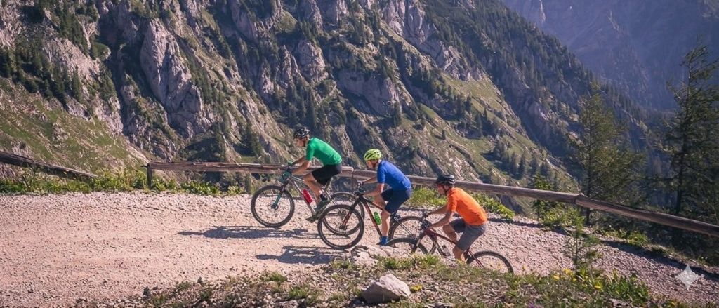 Drei Radfahrer fahren auf einem Bergpfad. Einer trägt ein grünes Shirt, einer ein blaues und der andere ein orangefarbenes Shirt. Der Weg ist von einem Holzgeländer flankiert.