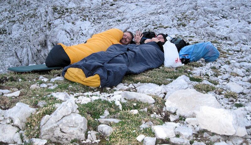 Zwei Personen ruhen auf einem felsigen Berg, einer in einem gelben Schlafsack und der andere in Blau, mit Taschen in der Nähe.