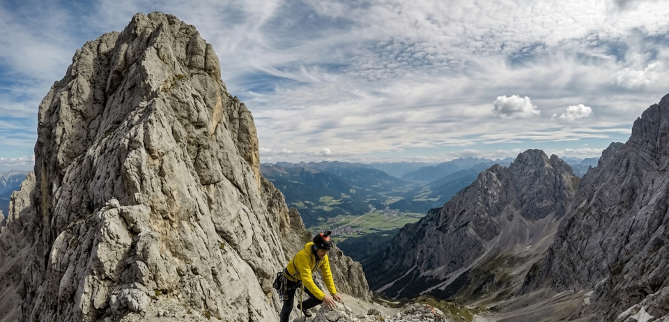 Ein Mann klettert einen felsigen Berghang hinauf, trägt eine gelbe Jacke und einen Helm. Unten ist das Tal mit einem Fluss und Häusern sichtbar.