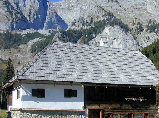 Ein traditionelles Holzhaus mit grauem Dach, in einem bergigen Gebiet. Das Haus hat einen Steinsockel und schwarze Fensterläden. Eine Person sitzt in der Nähe des Eingangs, mit Blumen und Grünpflanzen rund um das Haus.