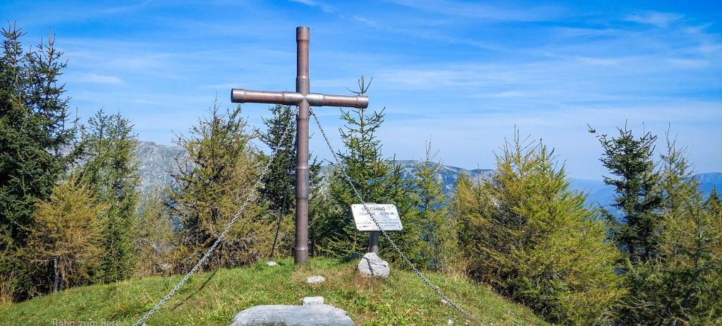 Ein Metallkreuz mit Ketten steht auf einem Berg in den Bergen. Es hat ein kleines Schild und ist von Kiefern umgeben. Der Himmel ist klar.
