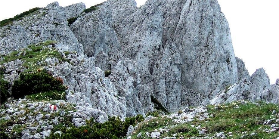 Ein zerklüfteter Berg mit steilen Felswänden und felsigen Vorsprüngen. Ein kleiner rot-weißer Flaggenaufsatz ist am Boden.