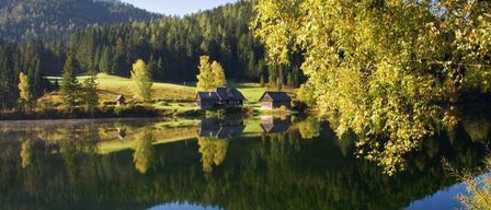 Eine friedliche Landschaft mit einem See, umgeben von üppigem Grün, Bergen in der Ferne und einigen rustikalen Häusern mit Spiegelungen auf dem Wasser.