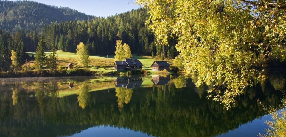 Eine friedliche Landschaft mit einem See, umgeben von üppigem Grün, Bergen in der Ferne und einigen rustikalen Häusern mit Spiegelungen auf dem Wasser.