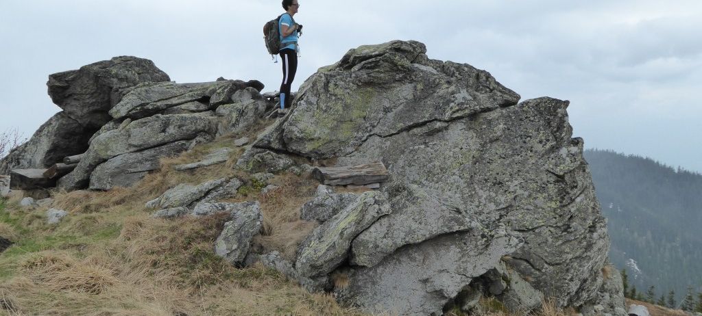 Ein Wanderer steht auf einem felsigen Vorsprung, trägt ein blaues Shirt und schwarze Hose. Der Wanderer hat einen Rucksack und scheint für ein Foto zu posieren.