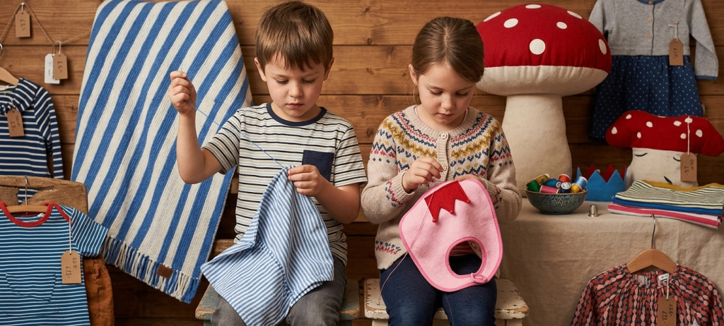 Zwei Kinder sitzen, eines hält ein blaues Tuch und das andere ein rosa Lätzchen, beide scheinen zu nähen. Hinter ihnen befindet sich eine Holzwand, die mit einem pilzförmigen Kissen verziert ist.