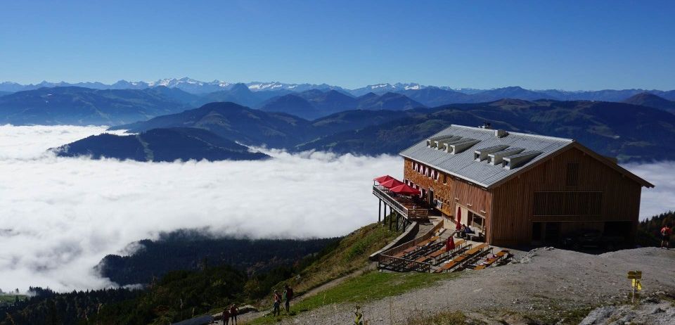 Eine Berghütte mit roten Schirmen steht auf einem Hügel, umgeben von grünem Gras und felsigem Gelände, mit einer malerischen Aussicht auf die Berge und Wolken.