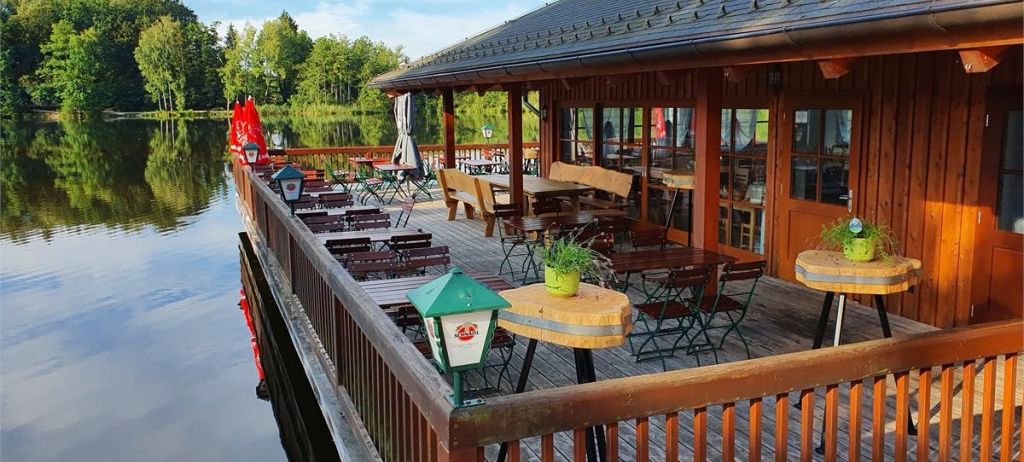 Ein Restaurant am Seeufer mit Holzbänken und Tischen, geschmückt mit grünen Pflanzen, und Blick auf einen ruhigen See und üppige Bäume.