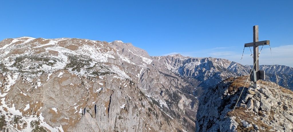 Ein atemberaubender Blick von einem Berggipfel, mit einem Kreuz, das den Gipfel markiert. Schneebedeckte Berge erstrecken sich in der Ferne unter einem klaren blauen Himmel.