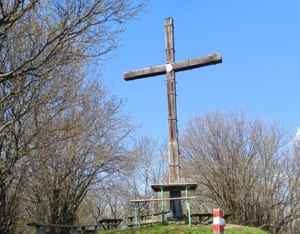 Ein hölzernes Kreuz steht auf einer grasbewachsenen Anhöhe, umgeben von kahlen Bäumen und Picknicktischen. Der Himmel ist klar und blau.