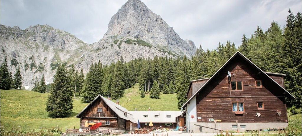 Zwei Holzchalets mit Bergkulisse. Bäume und ein grasbewachsenes Feld umgeben die Chalets. Ein roter Regenschirm steht vor einem Chalet.
