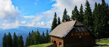 Ein kleines Holzhaus mit Solarmodulen auf dem Dach, eingebettet in einen Hügel, umgeben von grünen Bäumen und Bergen.