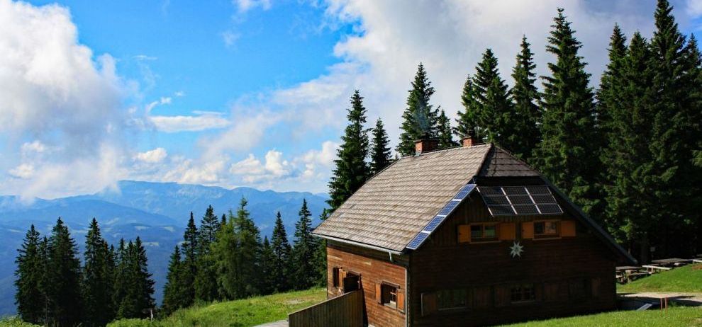 Ein kleines Holzhaus mit Solarmodulen auf dem Dach, eingebettet in einen Hügel, umgeben von grünen Bäumen und Bergen.