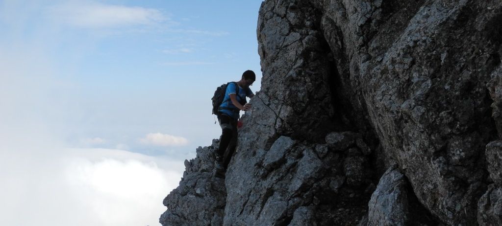 Ein Mann klettert einen steilen felsigen Felsen mit einem Rucksack und benutzt ein Metallwerkzeug zur Unterstützung.