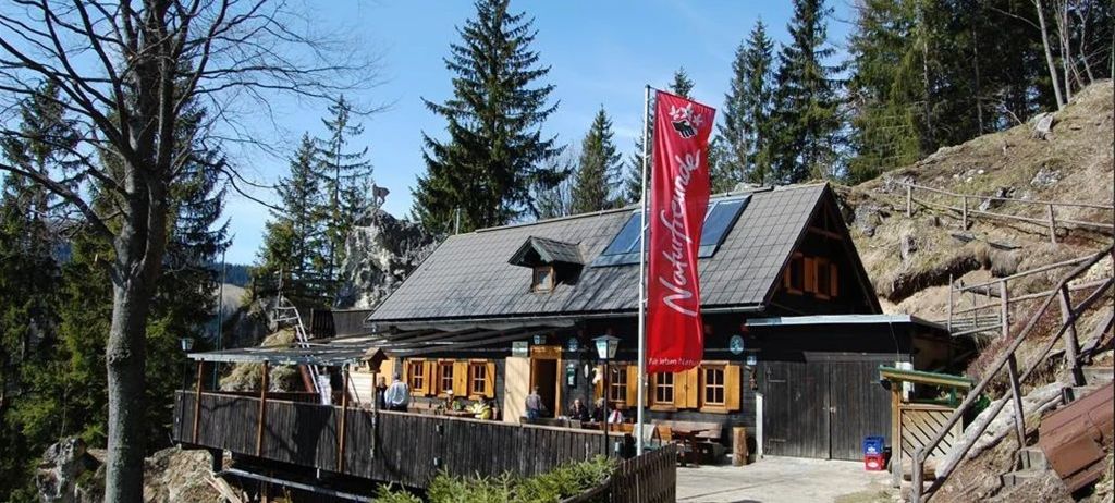Ein kleines, rustikales Chalet mit roter Fahne, Solarpanelen und überdachter Terrasse befindet sich an einem Berghang. Bäume umgeben das Chalet, und Menschen versammeln sich auf der Terrasse und im Inneren.