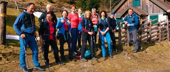 Eine Gruppe von Menschen, wahrscheinlich auf einer Wanderung, steht mit lächelnden Gesichtern zusammen. Sie tragen Wanderausrüstung und befinden sich in der Nähe eines Holzhauses und eines Zauns.