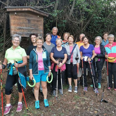 Eine Gruppe von Erwachsenen, alle mit Brille, posiert vor einem Schild mit der Aufschrift Romerbrucke. Sie befinden sich in einem Wald mit Bäumen und Pflanzen. Sie halten Trekkingstöcke in der Hand.