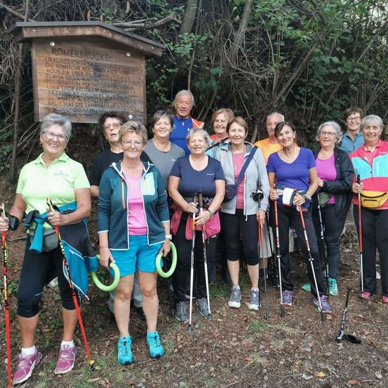 Eine Gruppe von Erwachsenen, alle mit Brille, posiert vor einem Schild mit der Aufschrift Romerbrucke. Sie befinden sich in einem Wald mit Bäumen und Pflanzen. Sie halten Trekkingstöcke in der Hand.