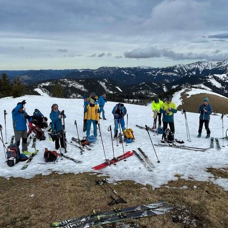 Eine Gruppe von Menschen, die in Skiausrüstung gekleidet sind, posiert mit ihren Skiern auf einem verschneiten Berggipfel unter bewölktem Himmel mit schneebedeckten Bergen in der Ferne.