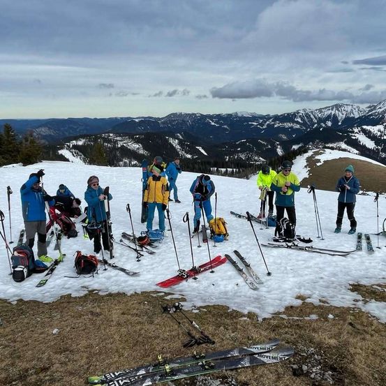 Eine Gruppe von Menschen, die in Skiausrüstung gekleidet sind, posiert mit ihren Skiern auf einem verschneiten Berggipfel unter bewölktem Himmel mit schneebedeckten Bergen in der Ferne.