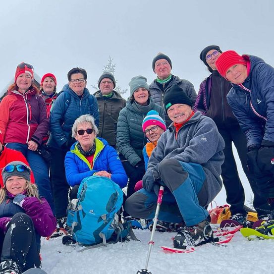 Eine Gruppe von Menschen, die in Winterkleidung gekleidet sind, posiert für ein Foto auf schneebedecktem Boden. Sie alle lächeln und einige halten Skistöcke.